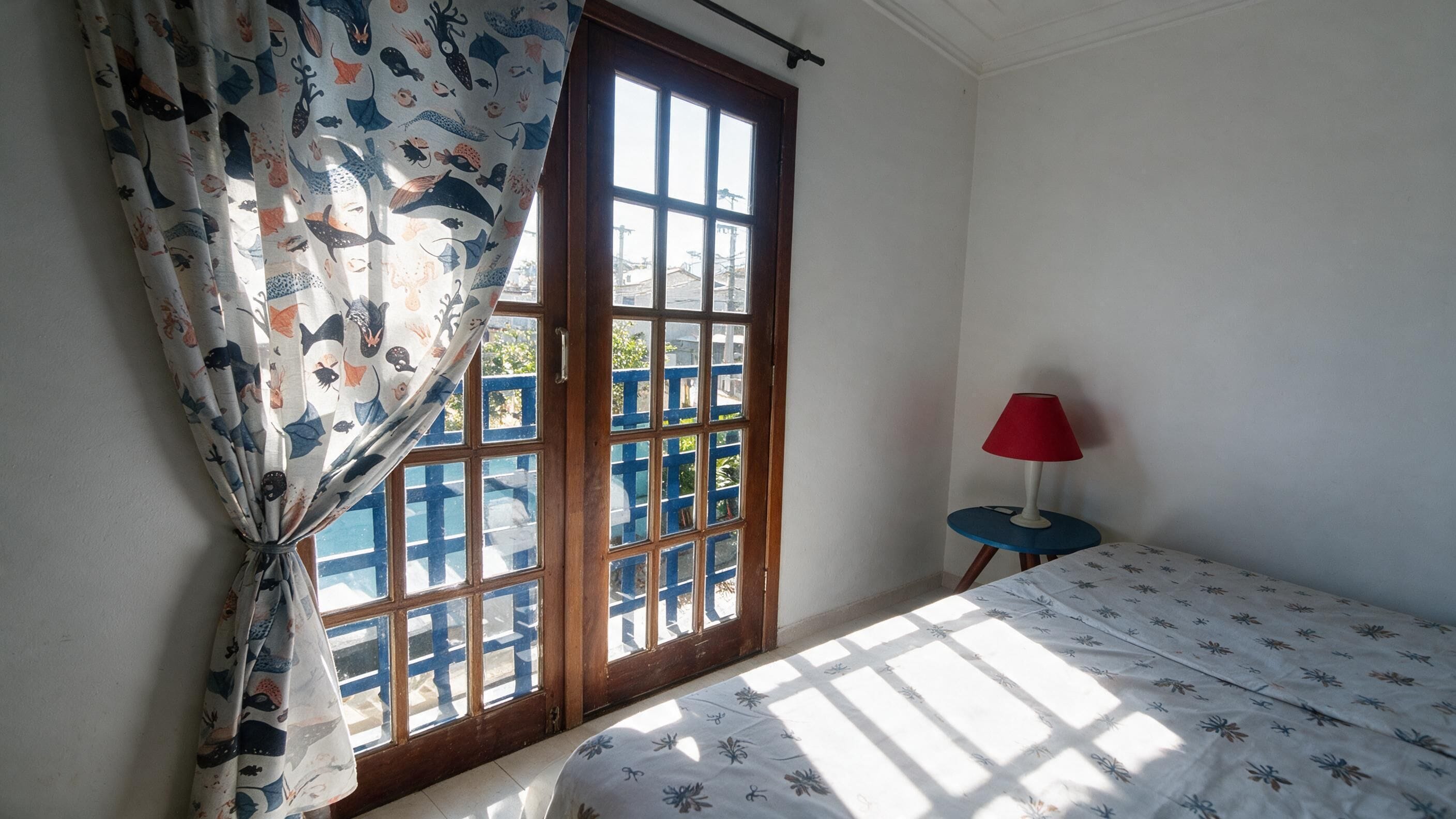 Een kamer met een bed, een raam met gordijnen, een klein tafeltje met een lamp en uitzicht op gebouwen.