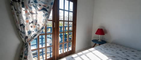 A room with a bed, a window with curtains, a small table with a lamp, and a view of buildings outside.