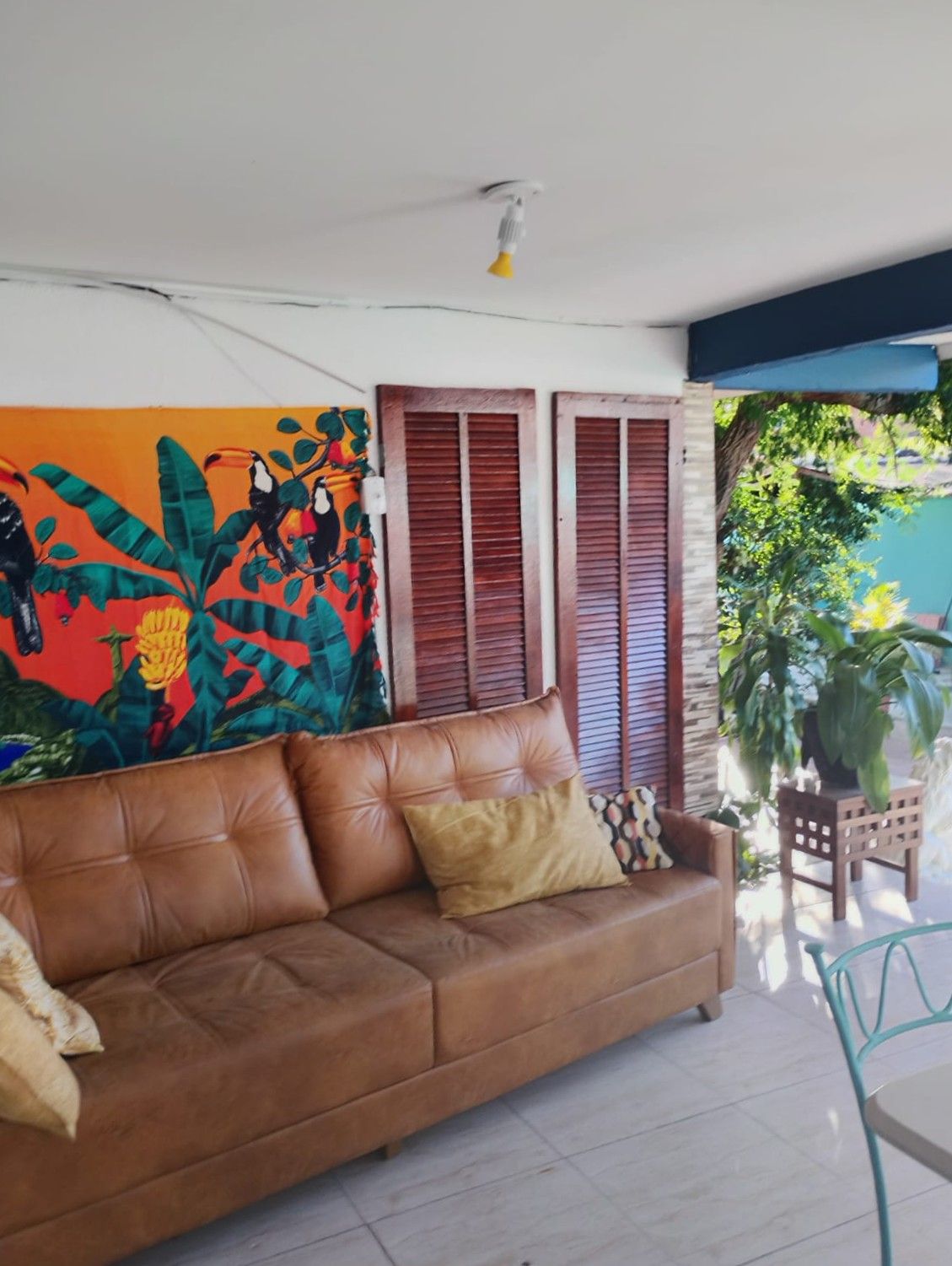 A room with a brown leather sofa, a colorful mural, wooden shutters, and a potted plant.
