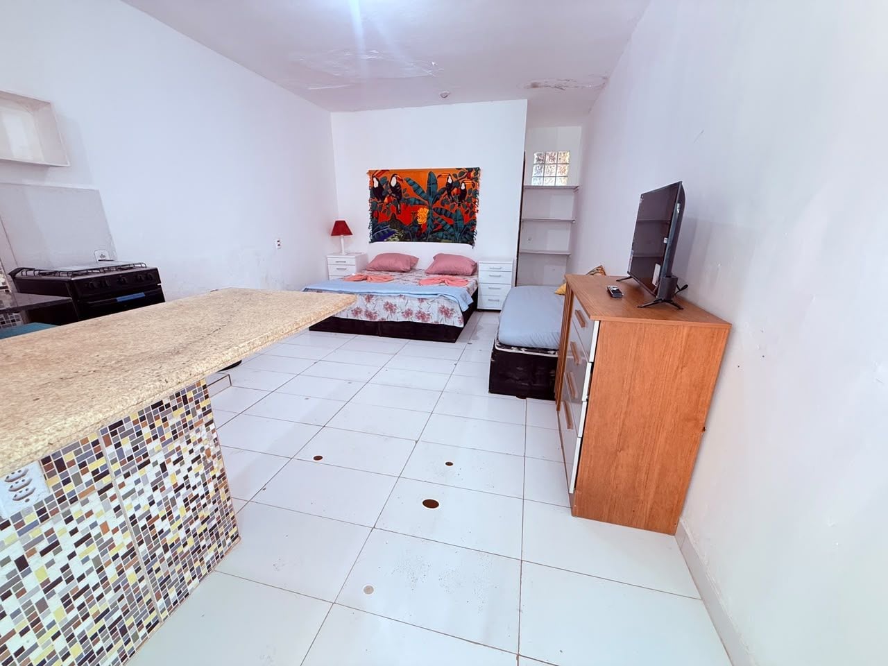 Una habitación compacta con kitchenette, una cama con cabecera colorida y un televisor sobre un mueble de madera.