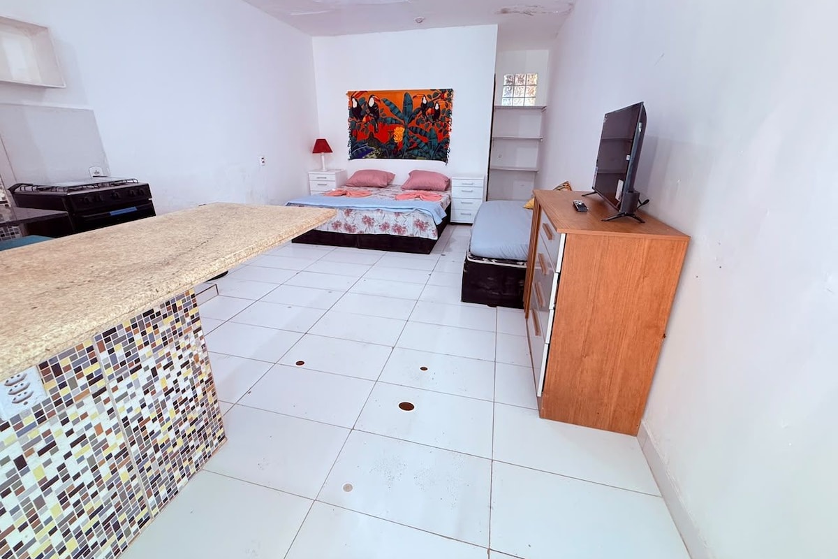 A compact room with a kitchenette, a bed with a colorful headboard, and a television on a wooden cabinet.