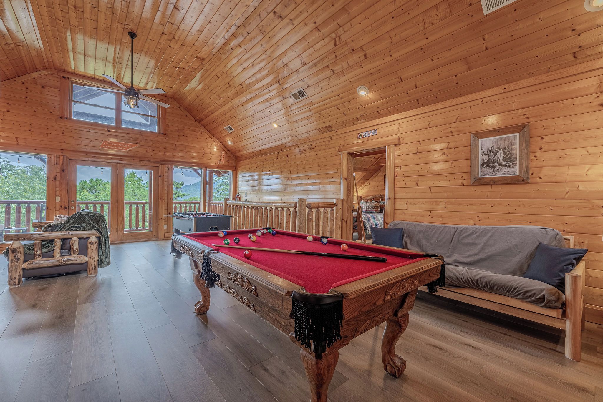 A wooden interior with a pool table, a sofa, and a view of the outdoors.