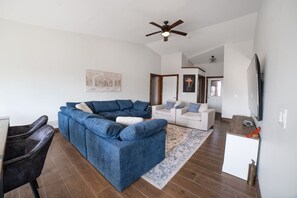Ein Wohnzimmer mit einem blauen Ecksofa, einem weißen Sofa, einem Fernseher und einem Deckenventilator.