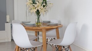 Un coin repas avec une table ronde en bois, dressée pour quatre personnes, des chaises blanches et un vase rempli de fleurs blanches.