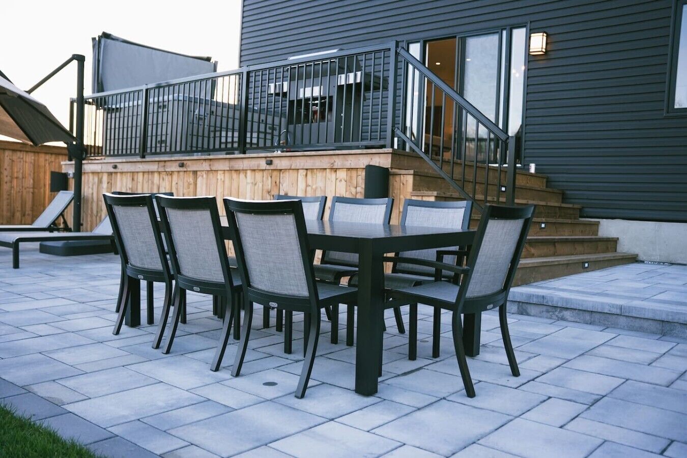 Un patio al aire libre con mesa y sillas, una terraza de madera y una escalera que lleva a un edificio de color oscuro.