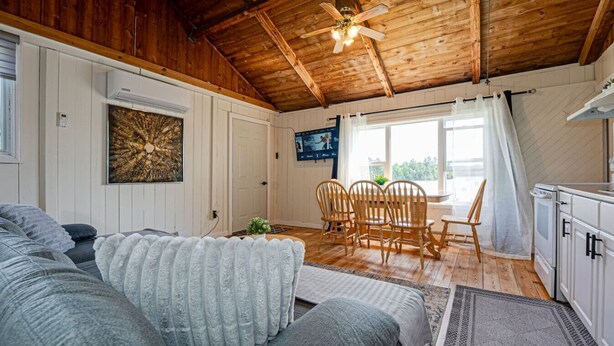 A cozy room with a wooden ceiling, a television mounted on the wall, a dining area with chairs, and a kitchen area with cabinets. - Beachfront Cottage #2- Maijac Resort & Marina (Burk`s Falls)