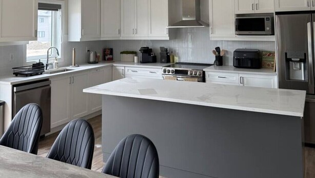 A modern kitchen with white cabinets, a central island, stainless steel appliances, and a dining area with chairs. - Modern Orillia Retreat - W/ Game Room (Severn)
