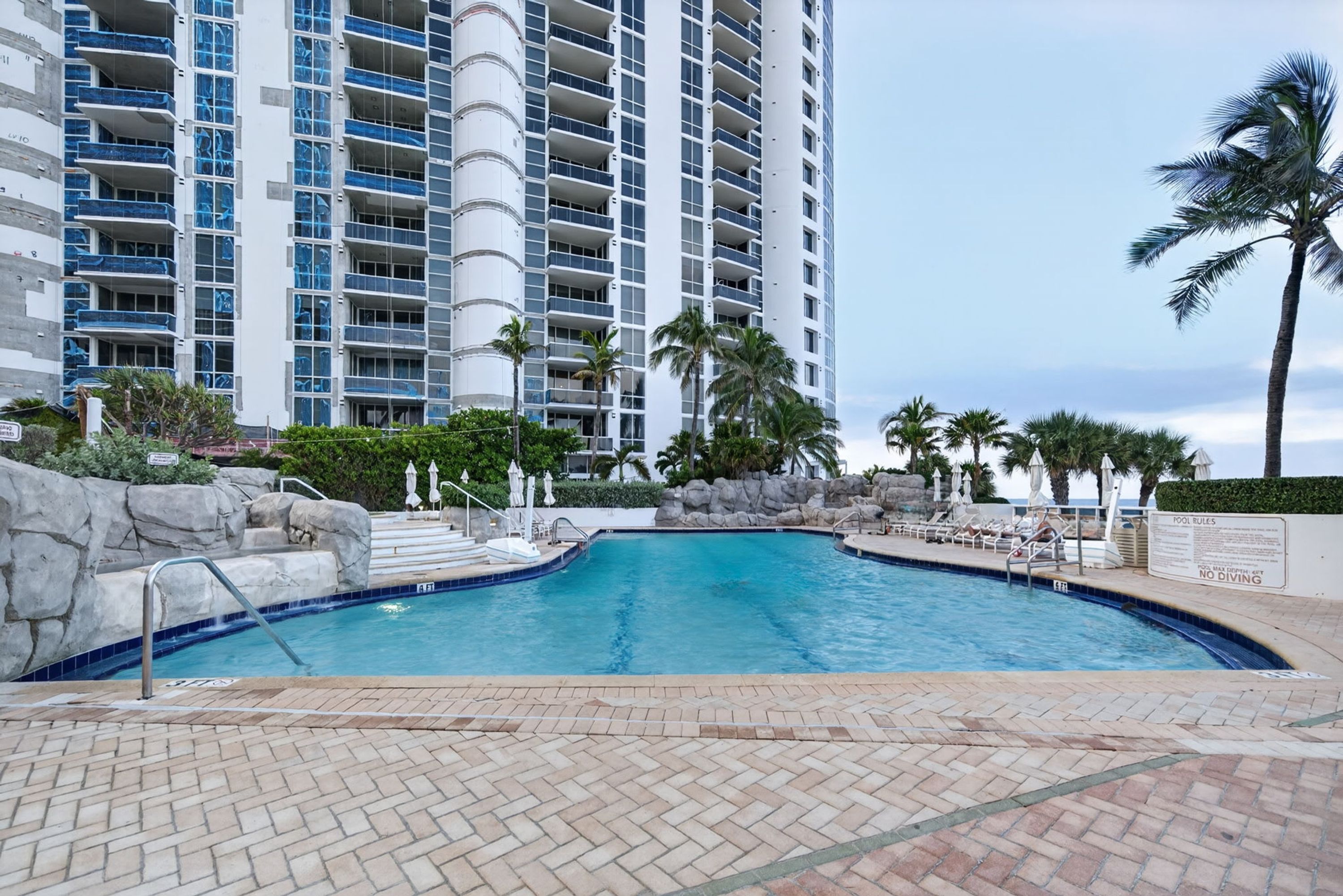 A swimming pool with a brick deck, surrounded by palm trees and a high-rise building in the background.