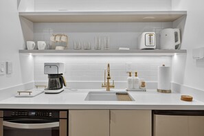 A modern kitchen with a built-in dishwasher, a coffee maker, a sink with a gold faucet, and a countertop with various kitchen items. - Luxury Beachfront Escape, Sunny Isles (Sunny Isles Beach)