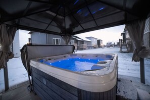Uma banheira de hidromassagem ao ar livre com área coberta, cercada por casas em um ambiente nevado.