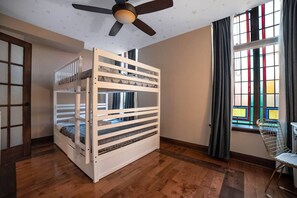 Una cama individual superpuesta en una habitación con ventilador de techo, ventana con cortinas y una silla.
