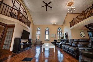 A spacious living room with a high ceiling, wooden staircase, and large windows.