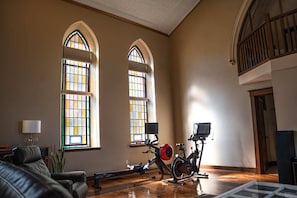 Amplia habitación con ventanas arqueadas, una bicicleta estática, un piano y una escalera.
