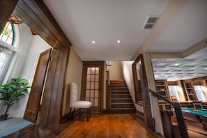 Una escalera de madera que conduce a un segundo piso, una silla blanca y una mesa de billar en una habitación con paneles de madera e iluminación empotrada en el techo.