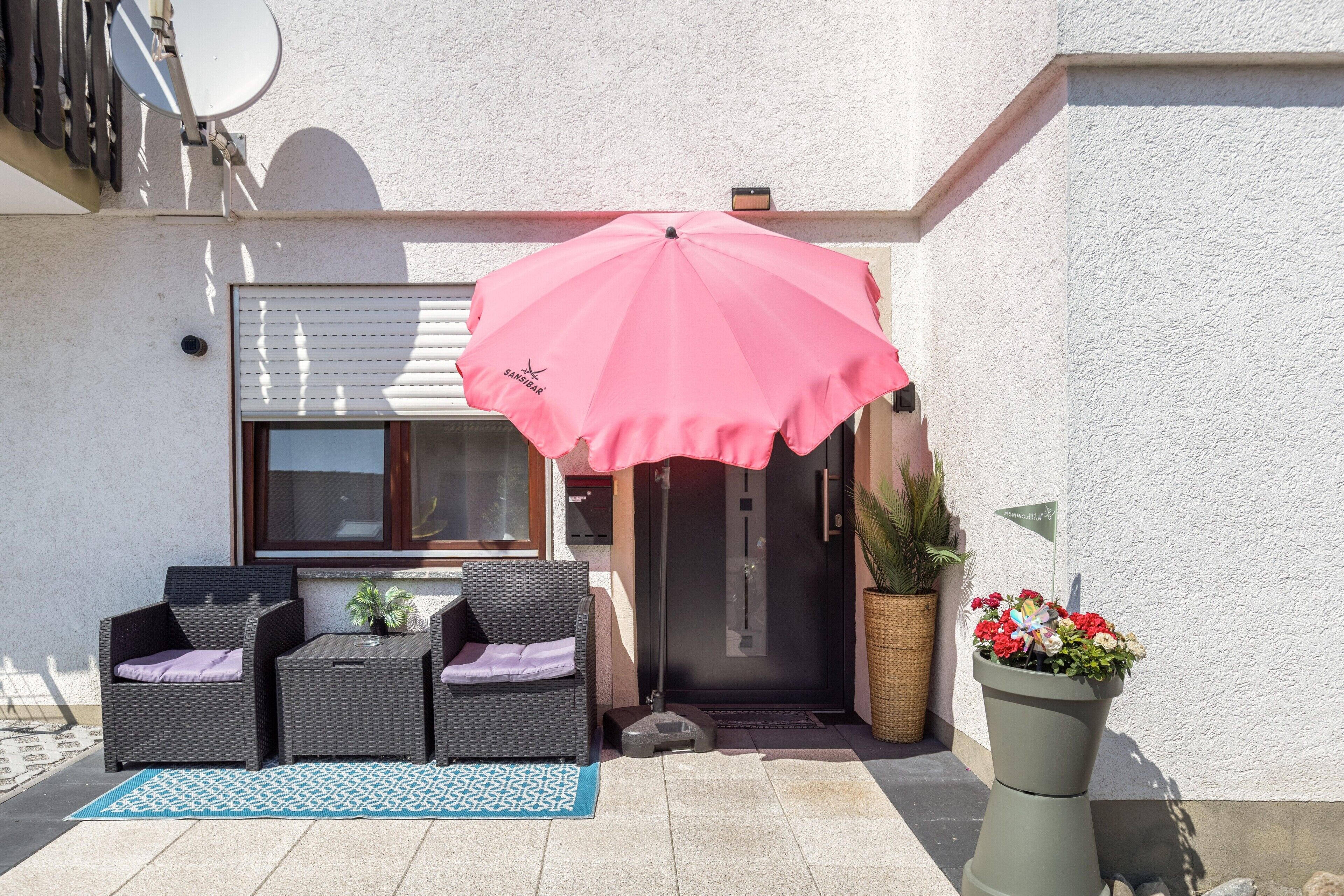 Ein Außenbereich mit Korbmöbeln, einem pinken Sonnenschirm und einem Blumentopf mit Blumen.