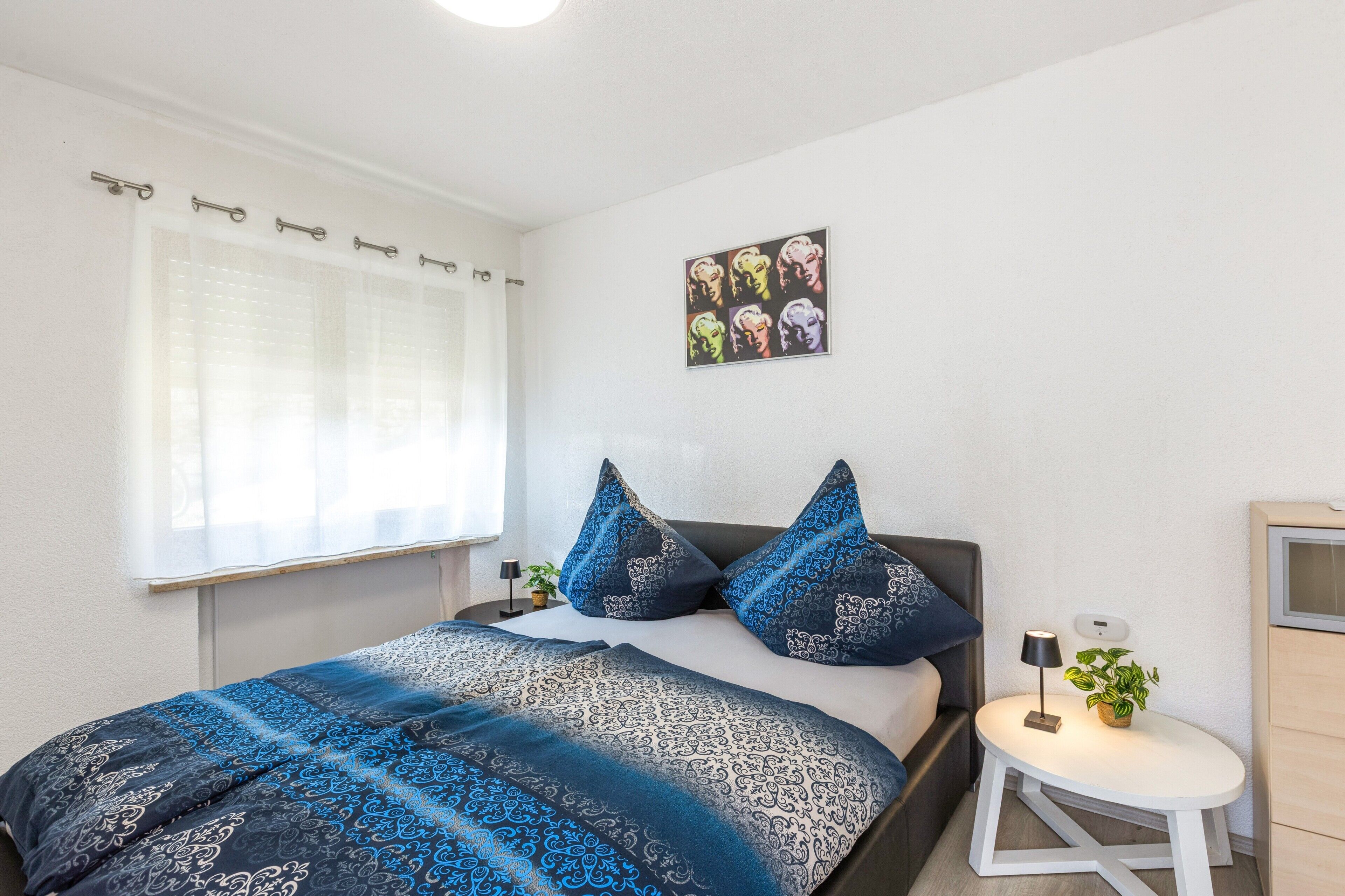 Ein Schlafzimmer mit einem Bett, Kissen, einem Nachttisch, einer Lampe, einer kleinen Pflanze und einem Wandbild.