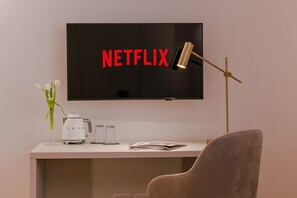 Ett modernt rum med en platt-TV som är monterad på väggen och visar ordet ”NETFLIX” med röda bokstäver, ett skrivbord med en kaffebryggare, två glas, en tidning, en lampa och en stol.
