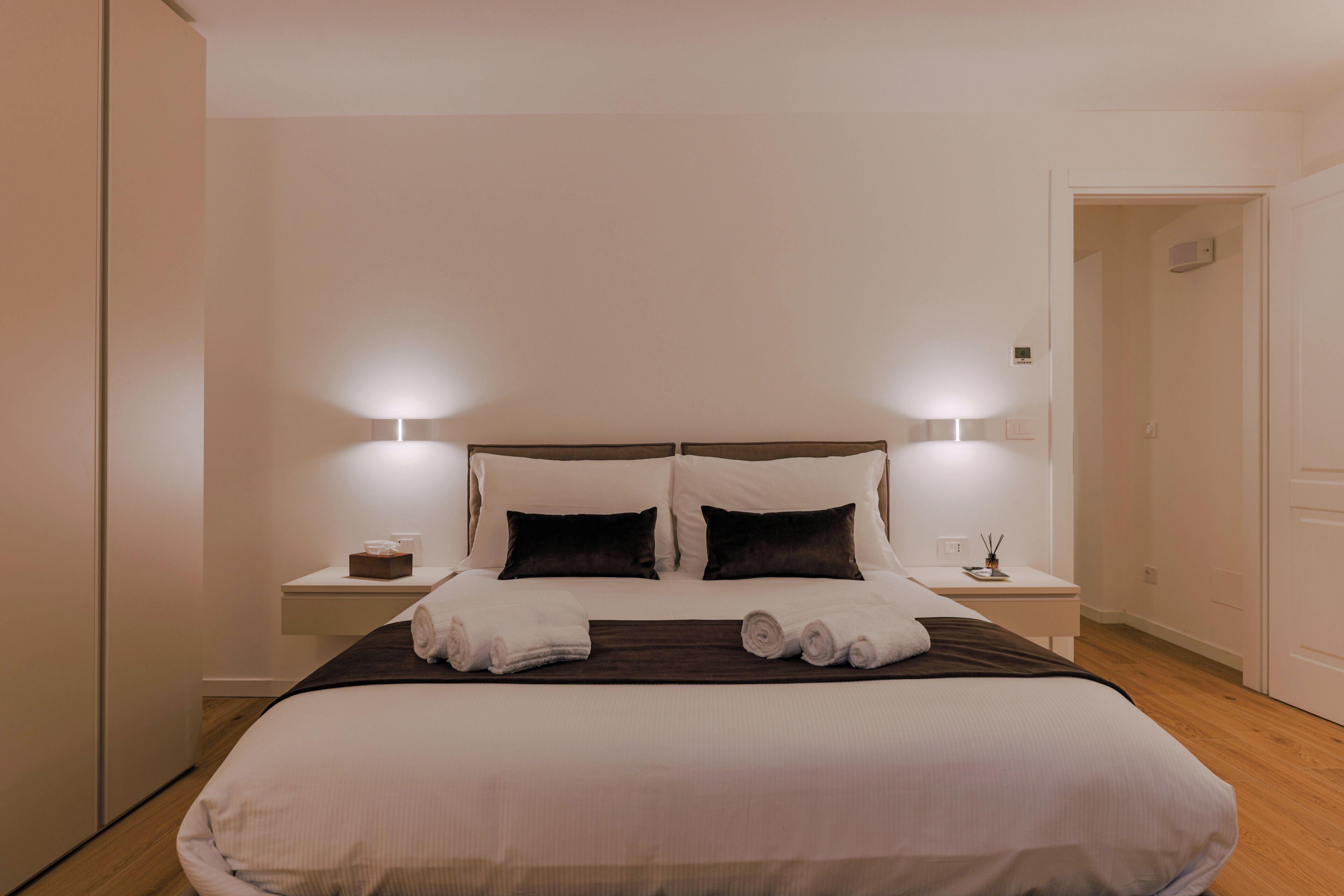 Ein ordentlich bezogenes Bett mit weißen Laken und zwei dunklen Kissen, ein Nachttisch mit einer Lampe sowie ein an der Wand befestigtes Thermostat.