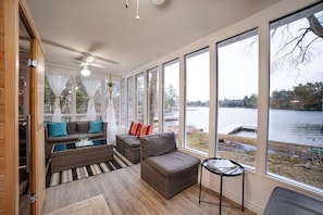 A room with a large window overlooking a lake, wicker furniture, and a ceiling fan. - Muskoka Waterfront Cottage - HOT TUB & SAUNA (Bala)