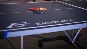 Taula de ping-pong amb una pilota i una paleta, una segona taula al fons, i un sostre amb llums fluorescents.