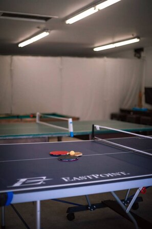 A ping pong table with a ball and paddle, a second table in the background, and a ceiling with fluorescent lighting.