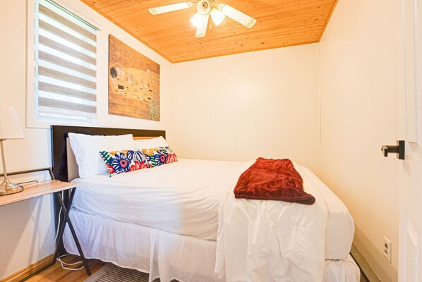 A bedroom with a bed, a ceiling fan, a window with blinds, a painting on the wall, and a nightstand with a lamp.