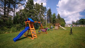 A playground with a slide, swings, and a climbing structure in a grassy area with trees and a building in the background. - Lake Cecebe Retreat #6- Maijac Resort & Marina (Burk`s Falls)