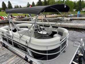 A pontoon boat docked at a pier with a covered seating area, a steering wheel, and a small table. - Burk`s Falls Beach #4 - Maijac Resort & Marina (Burk`s Falls)