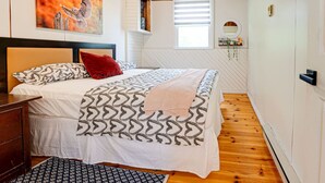 Un dormitorio con cama, cabecera, ventana con persianas, suelo de madera y un cuadro en la pared.