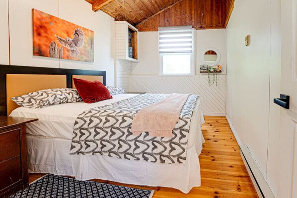 A bedroom with a bed, a headboard, a window with blinds, a wooden floor, and a painting on the wall.