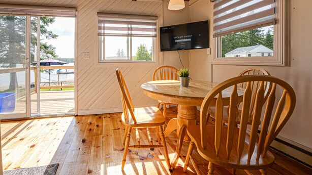 Un comedor de madera con una mesa y sillas, un televisor montado en la pared y ventanas amplias con persianas.