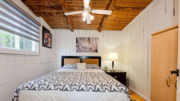 Un dormitorio con cama, ventilador de techo, mesita de noche con lámpara, puerta y ventana con persianas.