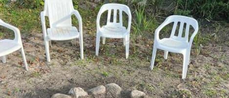 Un brasero con leña, rodeado por cuatro sillas de plástico blanco.