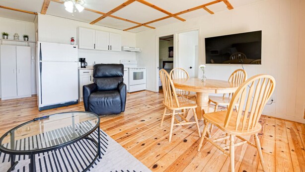A modern kitchen with white cabinets, a black leather chair, a dining table with wooden chairs, and a TV mounted on the wall. - Beachfront Cabin #5- Maijac Resort & Marina (Burk`s Falls)