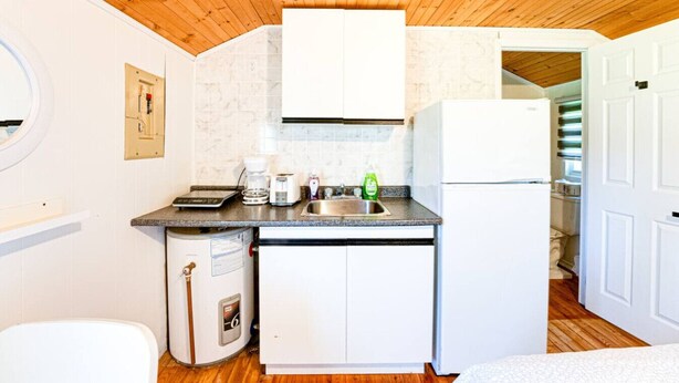 Ett kompakt kök med ett träpanelat tak, vita skåp, en diskho, en kaffebryggare och en diskmaskin.