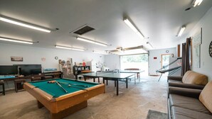 Ett biljardbord, ett bordtennisbord, en soffa och en tv i ett rum med ett betonggolv och vita väggar.