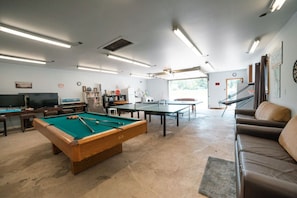 Ein Billardtisch, ein Tischtennistisch, eine Couch und ein Fernseher in einem Raum mit Betonboden und weißen Wänden.