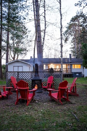 Un giardino sul retro con un camino a legna, poltrone Adirondack rosse e una casa sullo sfondo.
