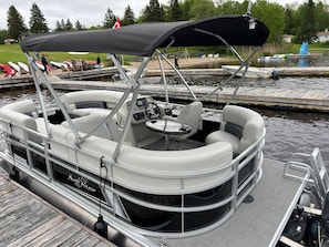 A pontoon boat docked at a marina with a covered seating area, a steering wheel, and a table.