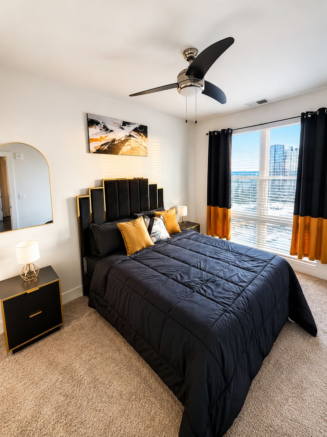 A modern bedroom with a large bed, a ceiling fan, a city view through the window, and a nightstand with a lamp.