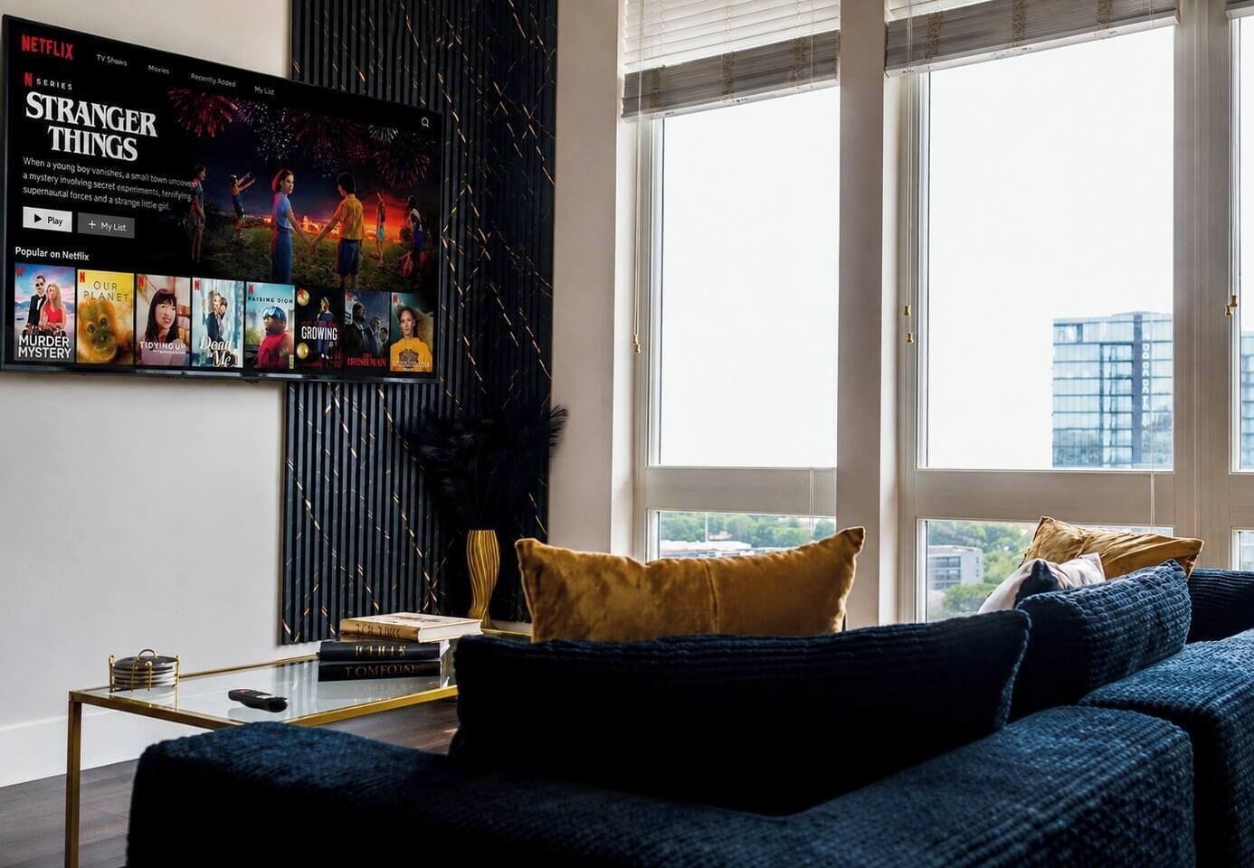 A living room with a large TV displaying a Netflix series, a sofa, a coffee table, and a window with a view.