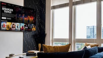 A living room with a large TV displaying a Netflix series, a sofa, a coffee table, and a window with a view.