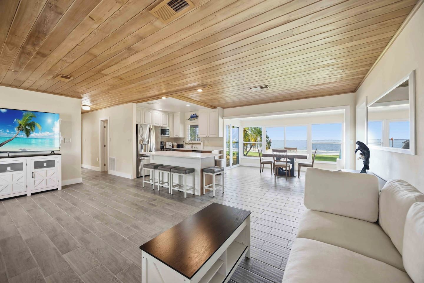 A spacious living area with a wooden ceiling, a kitchen with white cabinets, a dining area with a view, and a large window overlooking the ocean.