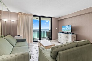 Ein Wohnzimmer mit einer Couch, einem Couchtisch, einem Fernseher auf einer Kommode und Blick auf den Ozean durch eine Schiebetür.