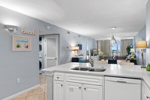 A modern kitchen with a built-in dishwasher, a sink, and a countertop. There is a dining area with a table and chairs, and a view of the ocean through the windows. - Gulf View| Private Beach| Pelican Terrace 1503 (Destin)