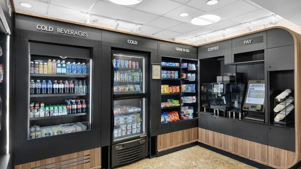 Una estación de autoservicio para bebidas con dispensadores refrigerados, una sección de productos para llevar y una zona de máquinas de café.