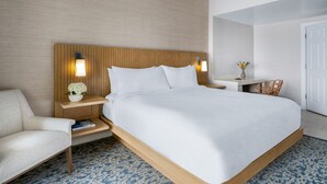 Habitación de hotel con una cama grande, mesitas de noche, una silla, una pequeña mesa con un florero y una puerta.