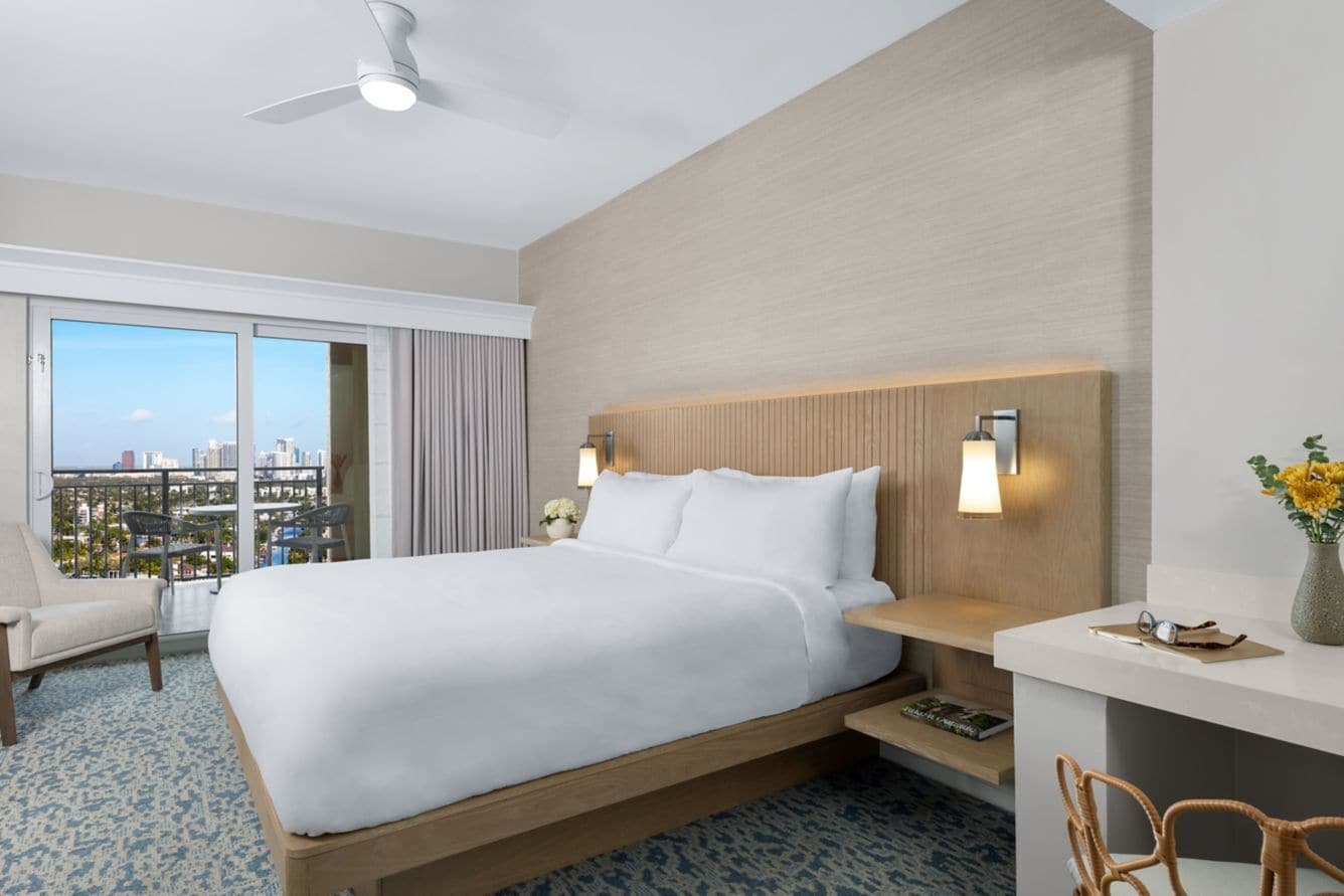 A hotel room with a bed, a desk, a chair, a ceiling fan, and a view of the city.