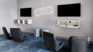 Ein Zimmer mit zwei wandmontierten Flachbildfernsehern, einer Spielkonsole und einem Regal mit einem Spiel-Controller. Vier schwarze Stühle mit blauen Kissen sind vorhanden.