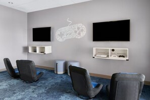 Quarto com duas televisões de tela plana fixadas na parede, um console de jogos e uma estante com um controle de videogame. Há quatro cadeiras pretas com almofadas azuis.